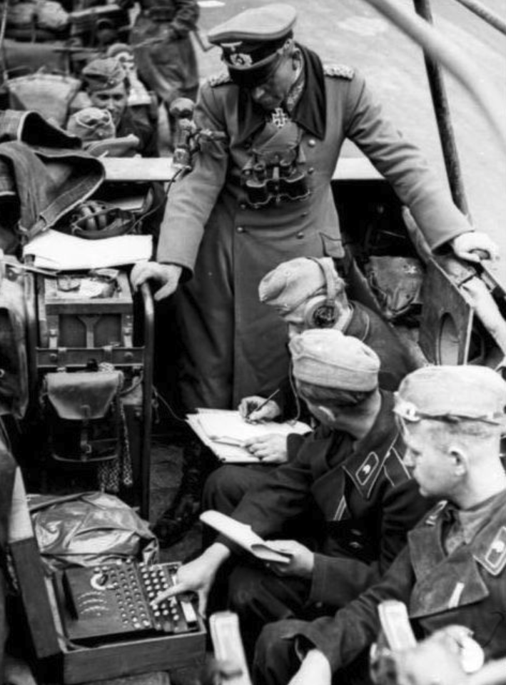 An officer watches soldiers operate an Enigma and write down a message inside a vehicle.
