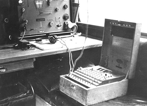 An open Enigma machine beside radio equipment and headphones.